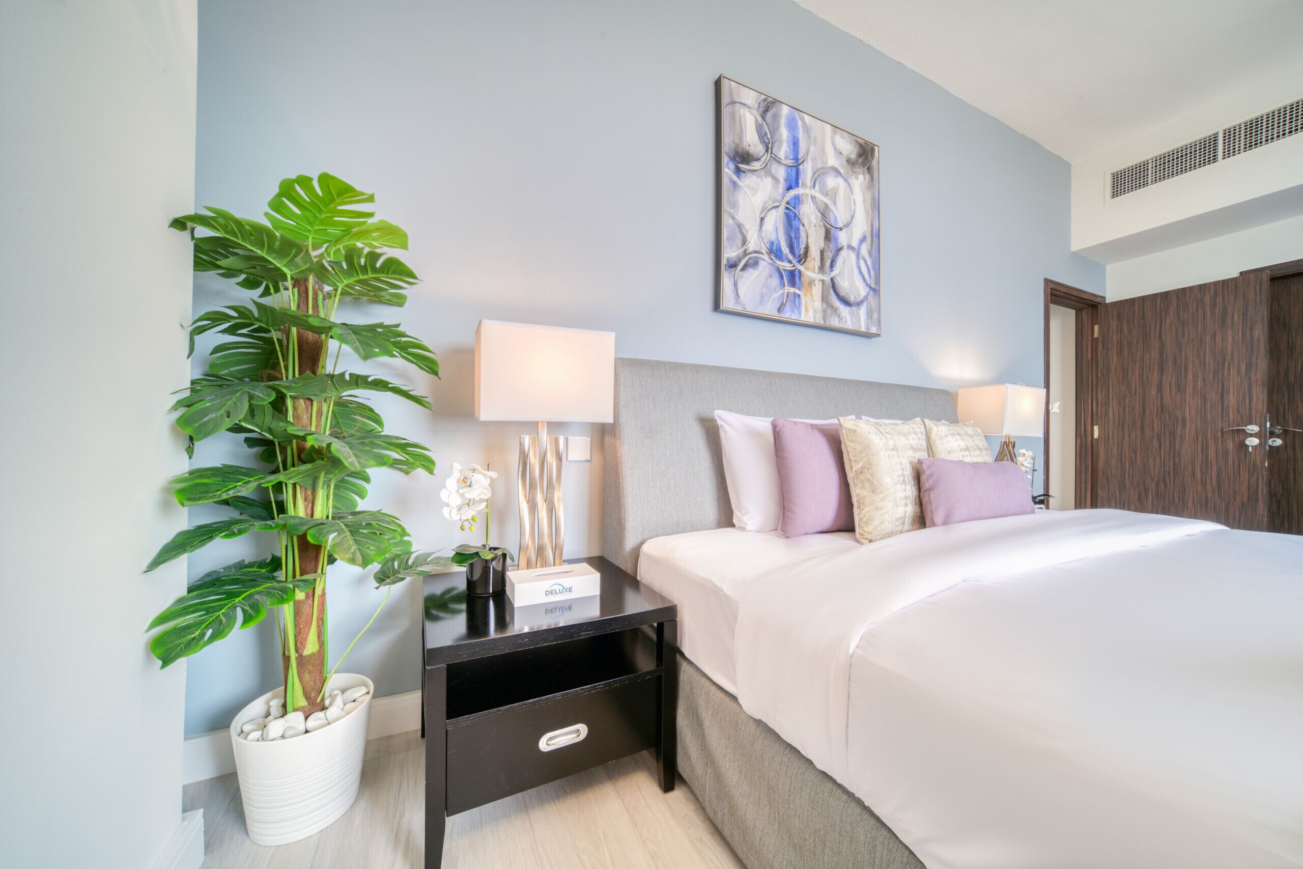 Een hotelkamer met een bed, nachtkastje, een bloempot, lamp en een schilderij aan de muur.