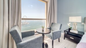 A hotel room with a large window, two armchairs, a small table, a bedside table with a lamp, and a bed.