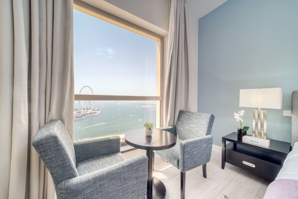 Quarto de hotel com uma janela grande, duas poltronas, uma mesa pequena, uma mesa de cabeceira com luminária e uma cama.