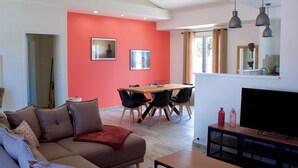 A modern living room with a red accent wall, a sofa, a dining table with chairs, and a flat-screen TV on a wooden cabinet. - Superbe Maison à 150m de la Plage (Saint-Pierre-d'Oléron)