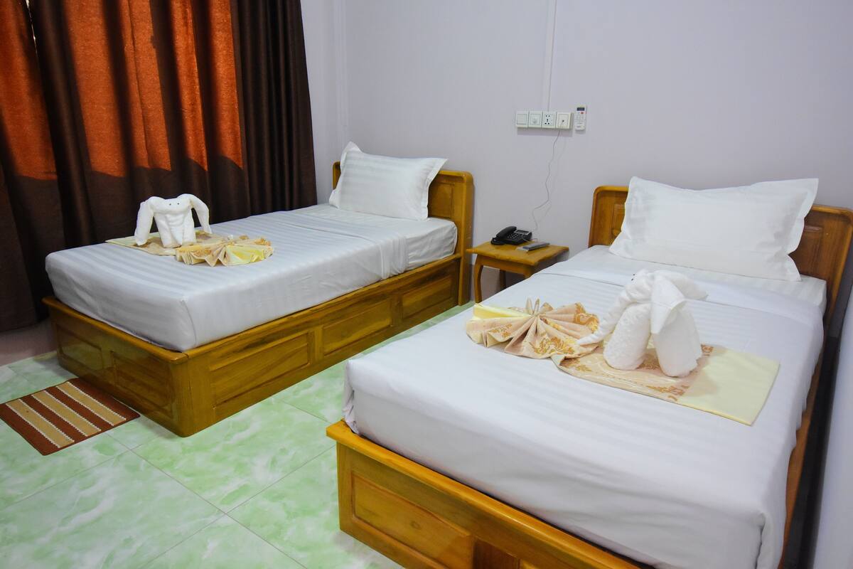 Two single beds with wooden frames and white linens, a bedside table with a phone, and a striped rug on a green-tiled floor.