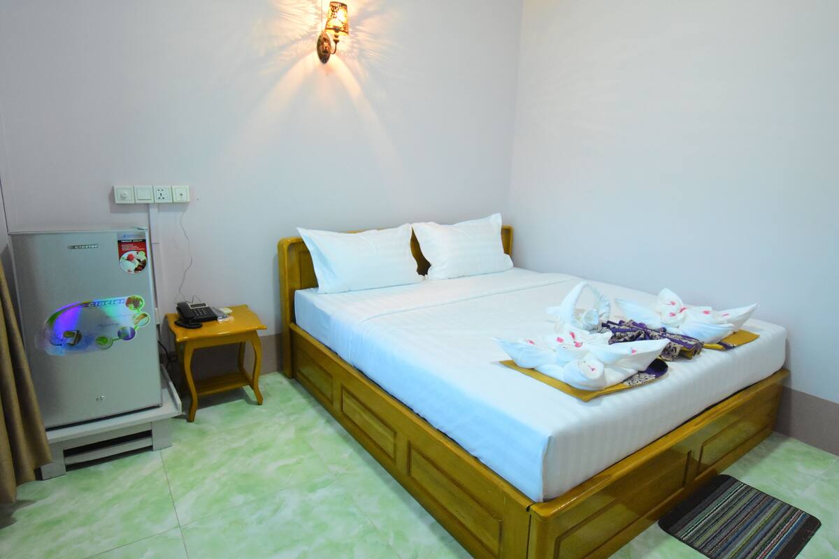 A hotel room with a bed, a small table, a refrigerator, and a wall-mounted light fixture.
