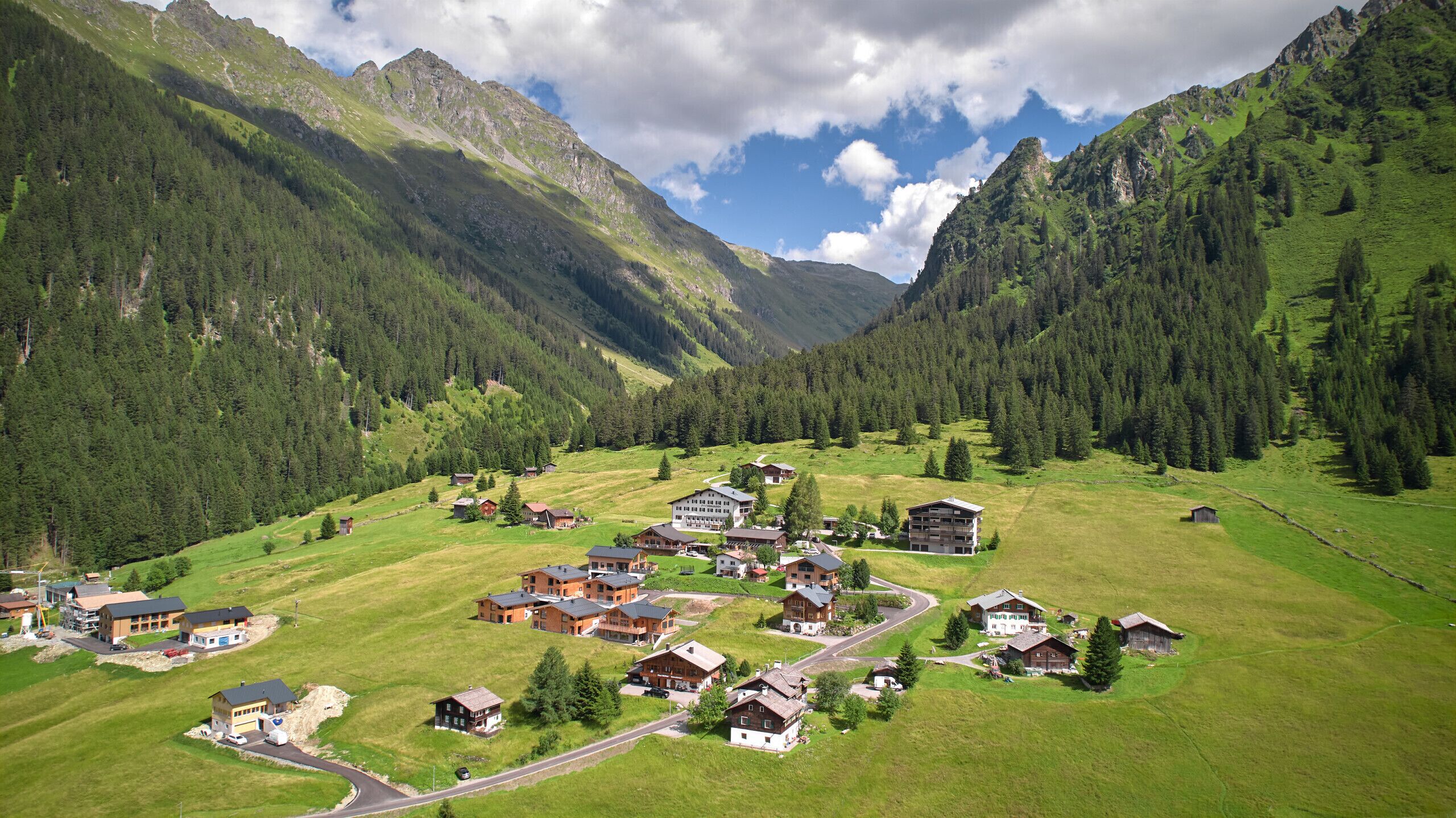 A mountain village with traditional wooden houses, a central building that appears to be a community center or hotel, and a road leading through the village.