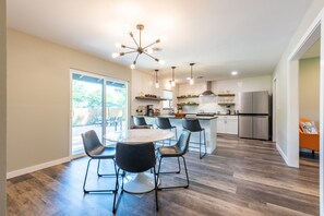 A modern kitchen with a dining area, featuring a central island, pendant lighting, and a large window overlooking a patio. - Zander Haus 3 Bedroom Home by RedAwning (Fredericksburg)