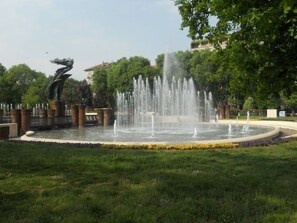 Stroll through a serene park setting surrounded by lush greenery and a central fountain with multiple water jets.