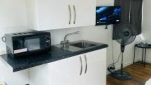 Een kleine keuken met witte kasten, een zwart aanrecht, een magnetron, een spoelbak, een ventilator en een televisie aan de muur.