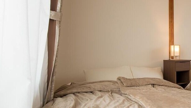 Een slaapkamer met een houten plafond, een bed met een beige dekbedovertrek, een nachtkastje met een lamp en een wit gordijn dat gedeeltelijk is opengetrokken.