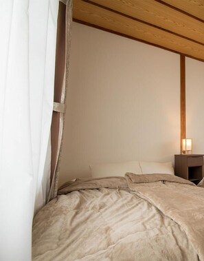 A bedroom with a wooden ceiling, a bed with a beige comforter, a nightstand with a lamp, and a white curtain partially drawn. - 7/// (Matsuyama)