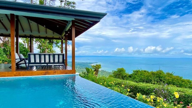 Una piscina infinita con vista al océano, un área de estar cubierta y exuberante vegetación.