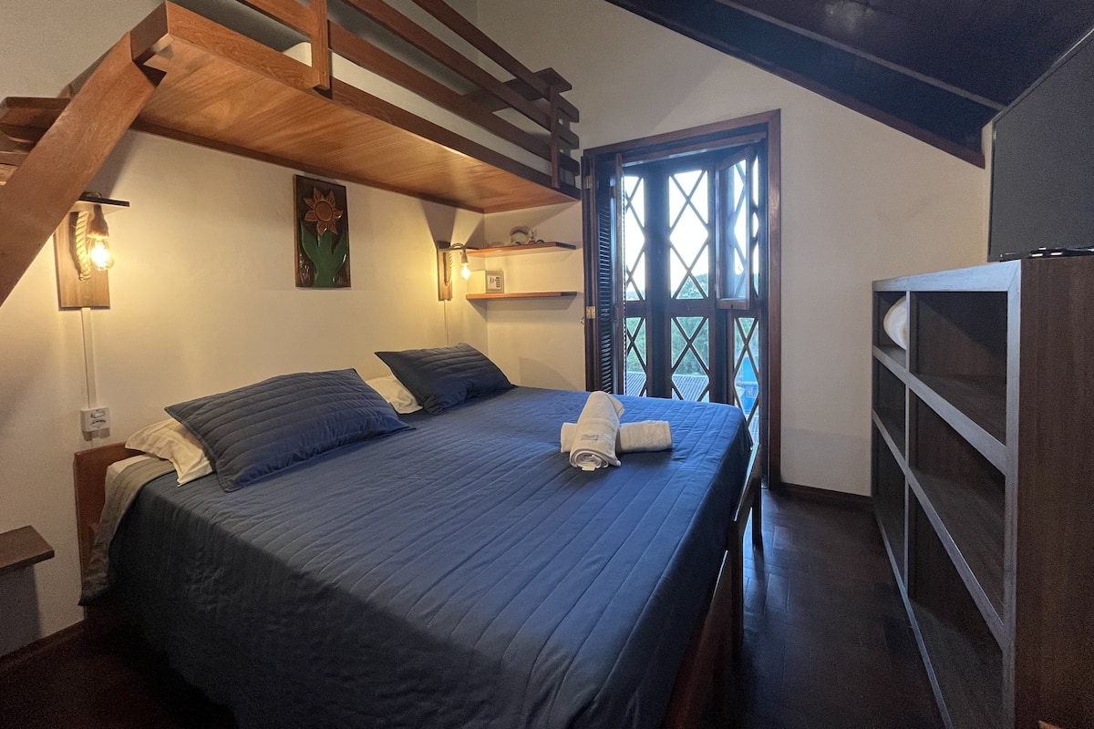 A bedroom with a bed, wooden ceiling beams, a window with a view, and a nightstand with a lamp.