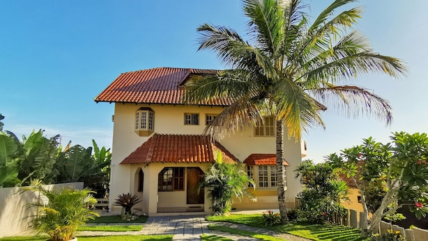 A two-story house with a tiled roof, a central entrance, and a well-maintained garden with palm trees and other tropical plants. - Pousada Canajurê (Florianópolis)