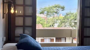 A bedroom with a bed, a window with a view of buildings and trees, and a wall-mounted light fixture. - Pousada Canajurê (Florianópolis)