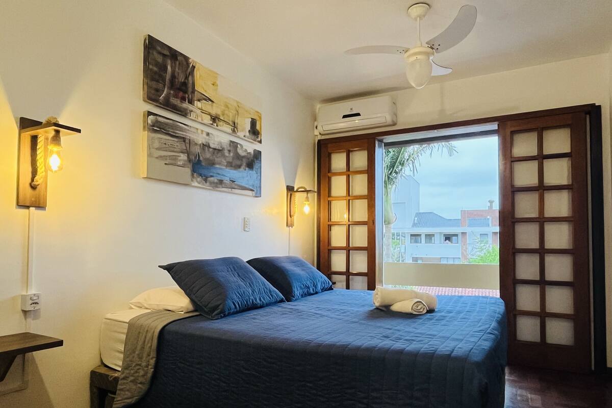 A bedroom with a bed, a ceiling fan, wall-mounted lights, and a window with a view of buildings.