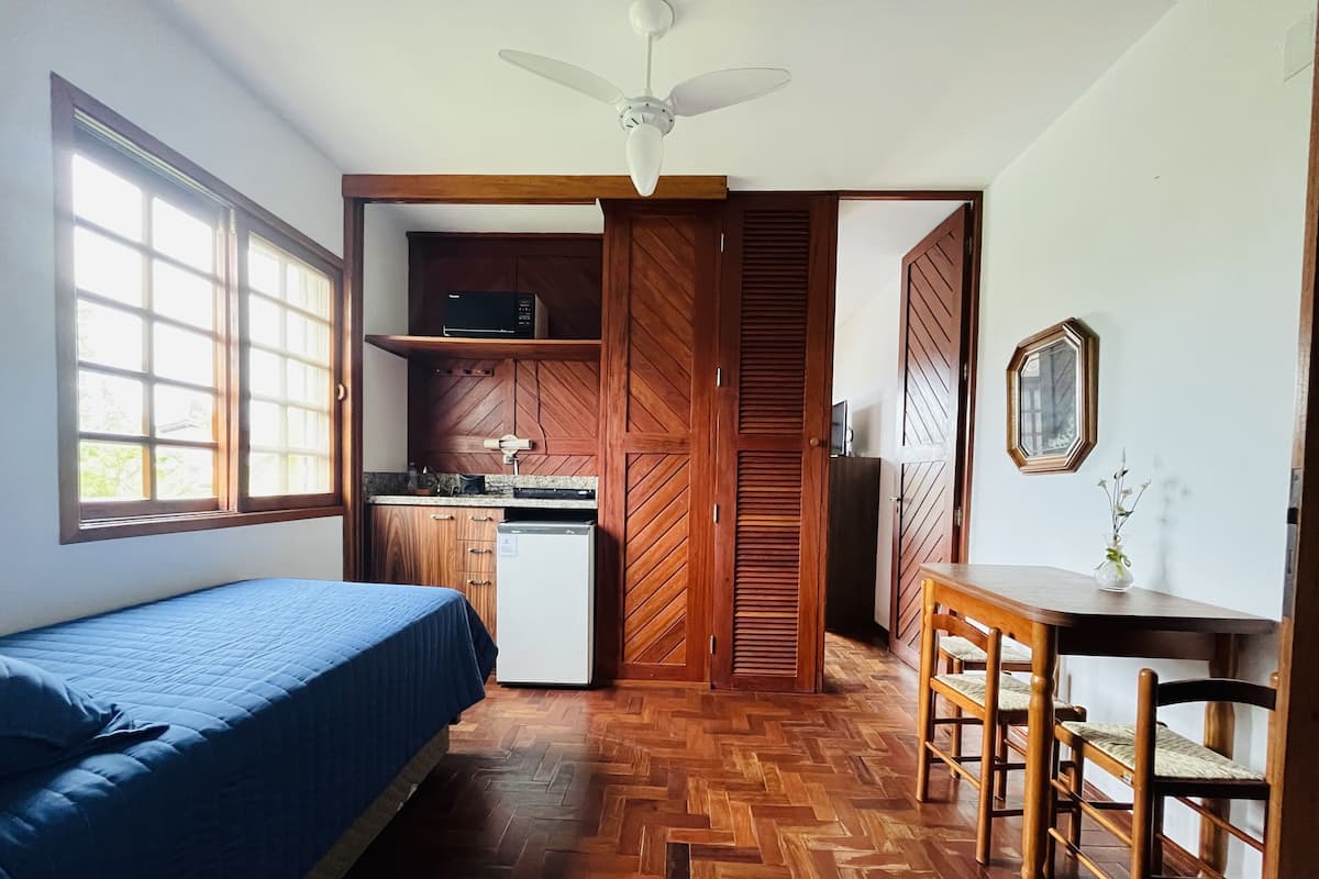 A compact room with a bed, a small kitchenette, a wooden wardrobe, a dining table with chairs, and a ceiling fan.