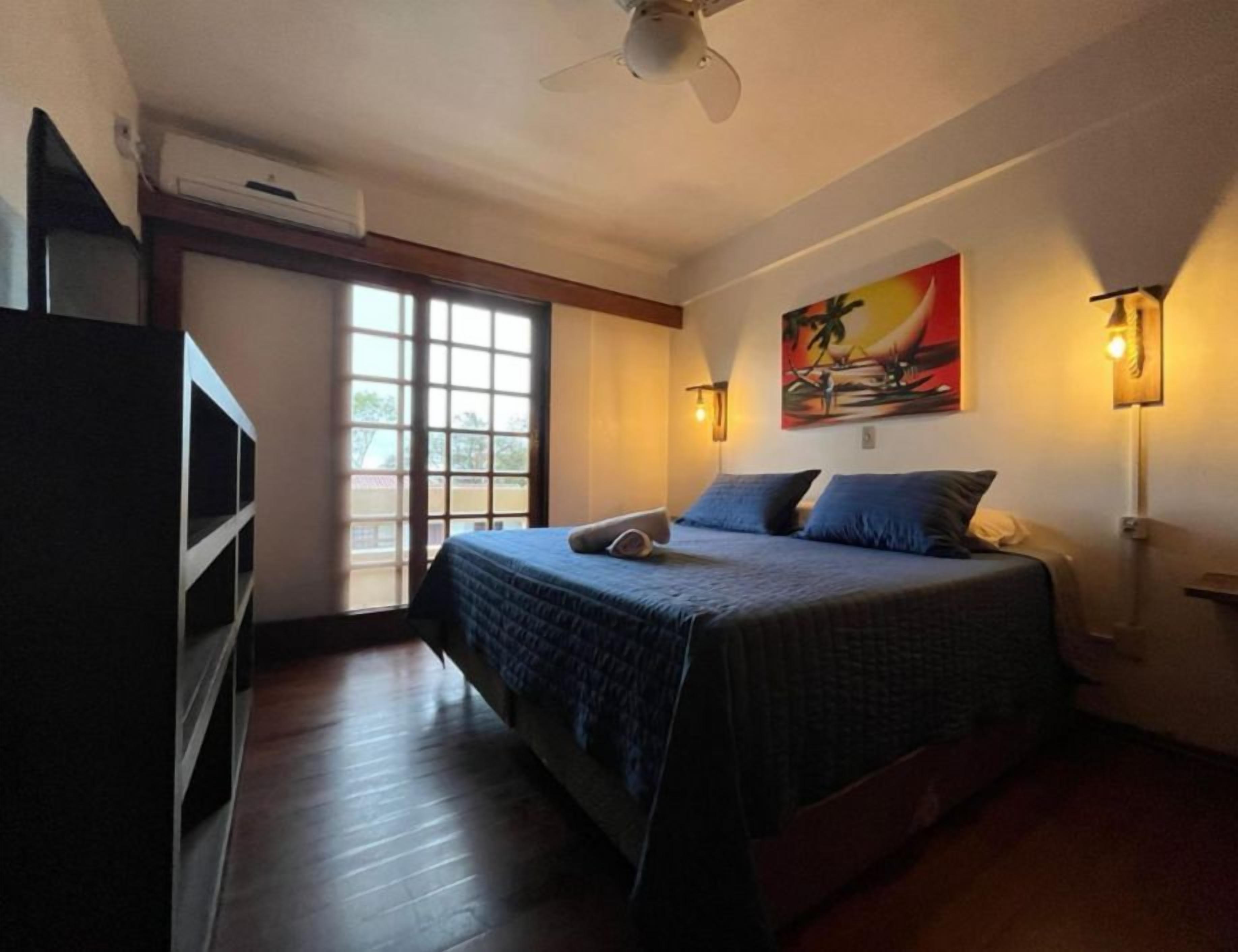Quarto com cama, janela, ventilador de teto e luminárias fixadas na parede.