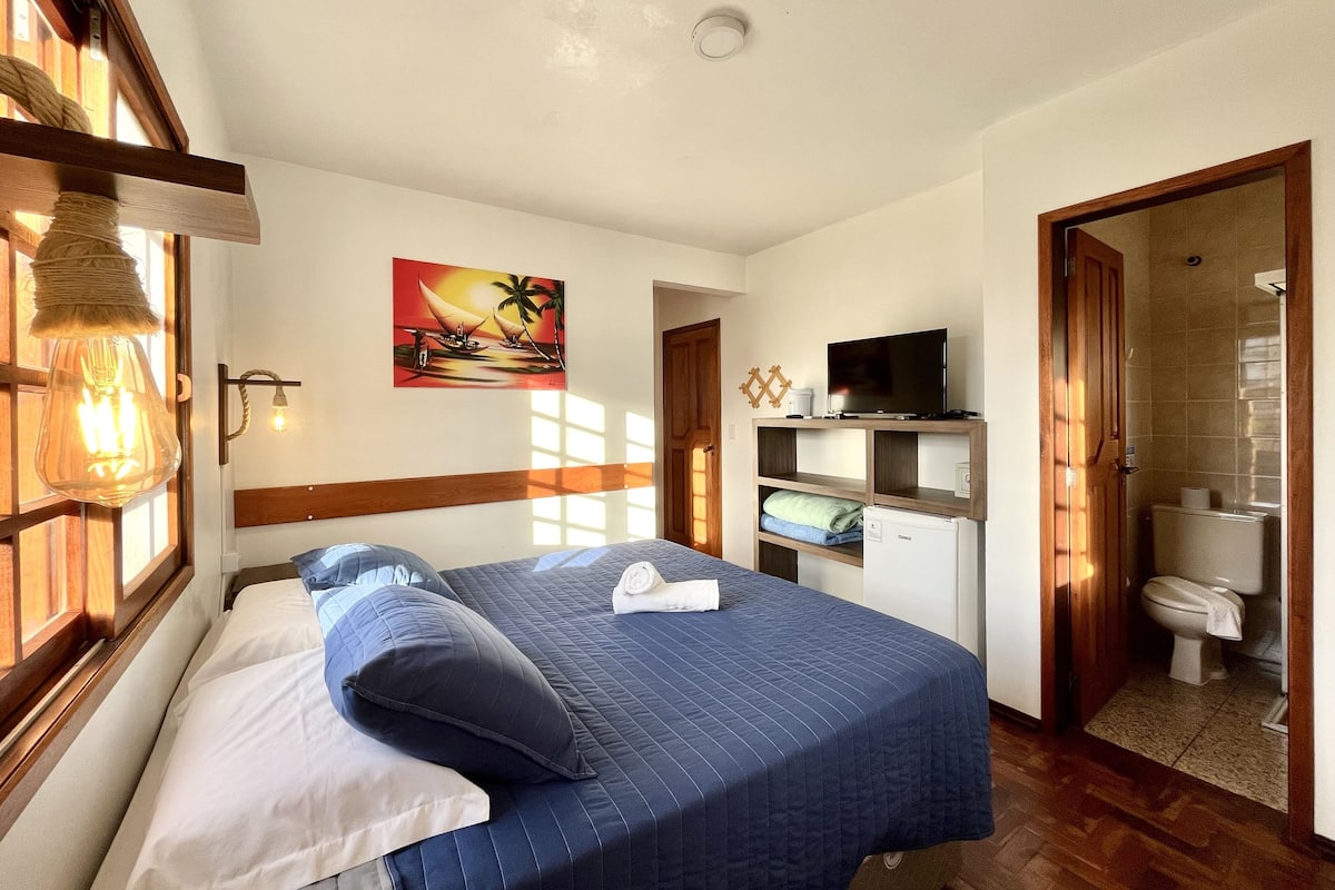 A bedroom with a bed, a television, a painting, and a bathroom visible through an open door.