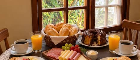 Et morgenbord dækket med forskellig mad, et vindue med blondegardiner og udsigt til grøn natur.