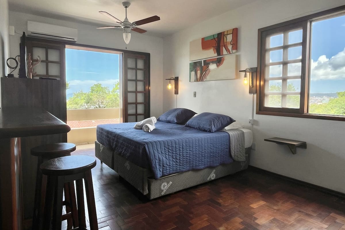 A bedroom with a bed, a ceiling fan, a window with a view, and a bar area.
