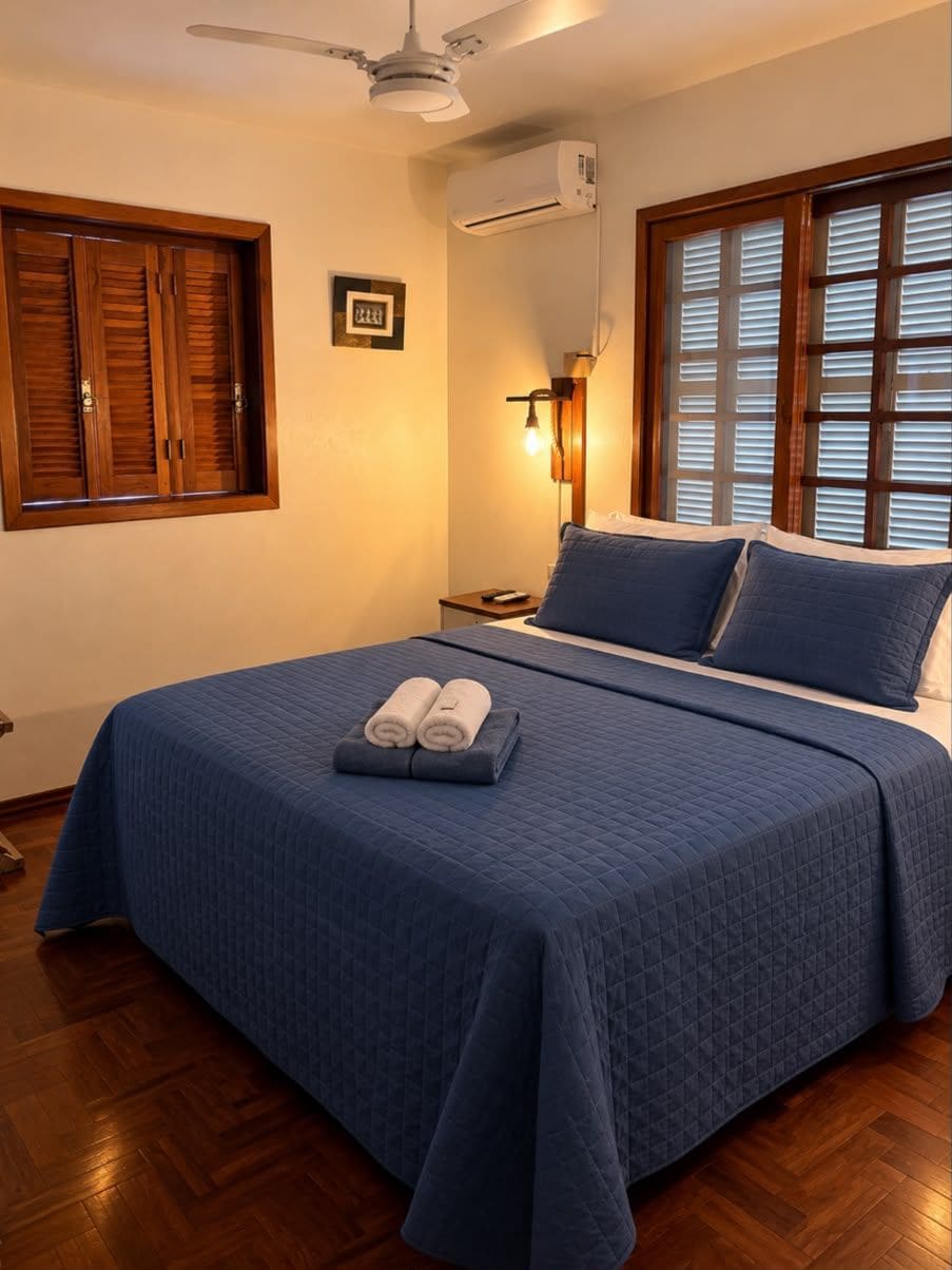 Une chambre à coucher comprenant un lit, un ventilateur de plafond, un appareil de climatisation, une table de chevet et une fenêtre avec des stores.