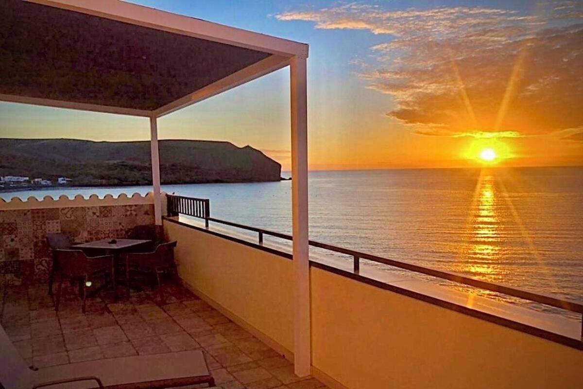 Un balcón con vista a la puesta de sol sobre el mar, una mesa y sillas.