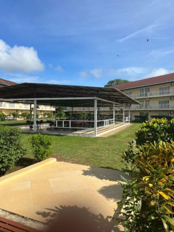 A covered outdoor area with a tiled floor, surrounded by greenery and a building with a red roof in the background. - Lungi Gateway Hotel (Lungi)