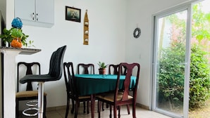 Un coin repas avec une table dressée pour quatre personnes, entourée de chaises en bois. On aperçoit une plante en pot sur la table, et un meuble avec deux portes au fond.