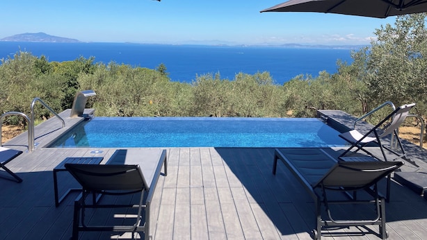Eine Holzterrasse mit Pool und Liegestühlen, mit Blick auf eine malerische Meereslandschaft und ferne Berge.