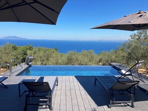 Une terrasse en bois avec une piscine et des chaises longues, offrant une vue panoramique sur la mer et les montagnes lointaines.