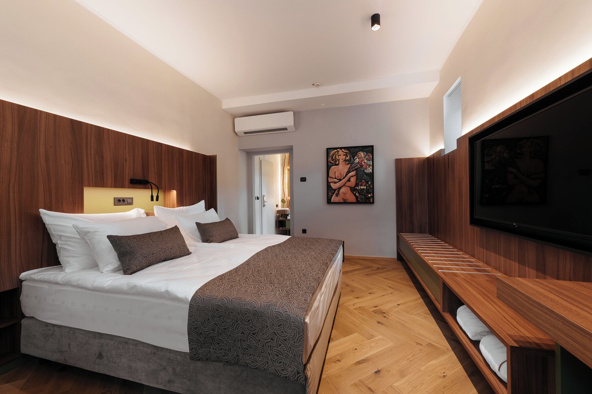A modern bedroom with a large bed, wooden paneling, a flat-screen TV, and a bench with towels.