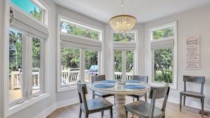 Un coin repas avec une table ronde, six chaises et un lustre. De grandes fenêtres offrent une vue sur l’extérieur.