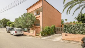 Un immeuble de deux étages avec un balcon à l’étage supérieur. Des voitures sont garées devant, et un portail mène à l’entrée.