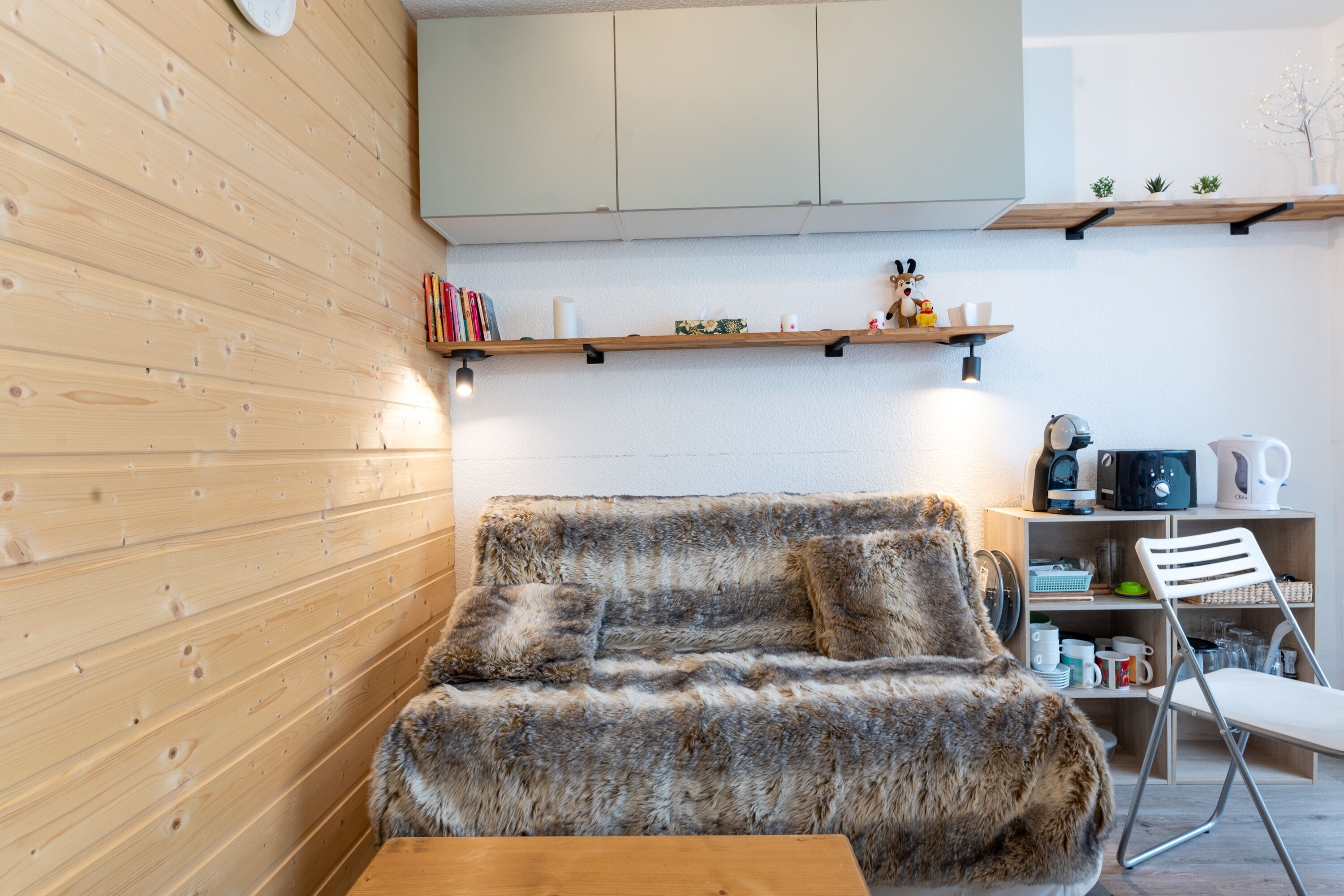 Ein gemütliches Zimmer mit Holzwand, einer Couch, einem kleinen Tisch und einer Küchenzeile mit Regalen und Geräten.