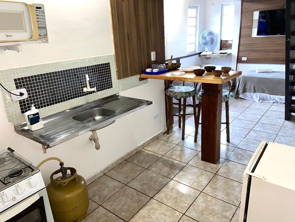 A compact kitchen with a gas stove, a sink, and a microwave. There is a dining area with a table and chairs, and a bed in the background. - MARÉ ALTA SUITES (São Sebastião)