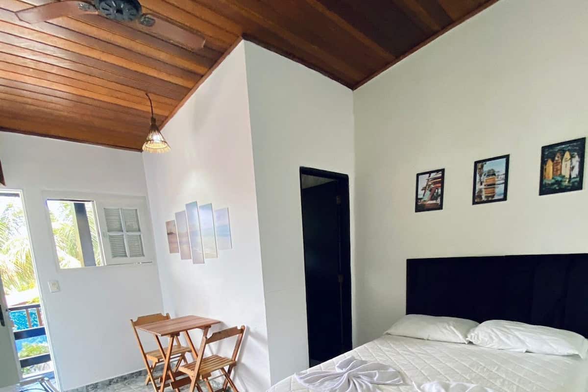 A bedroom with a bed, a dining table, and chairs, a ceiling fan, and a view of the outside.