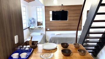 Quarto compacto com cama, televisão, ventoinha e área de refeições com tigelas e pratos.