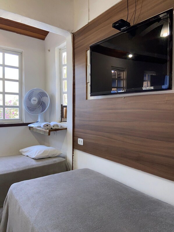 Un dormitorio con una cama, una mesita de noche con un ventilador, una ventana y un espejo en la pared.