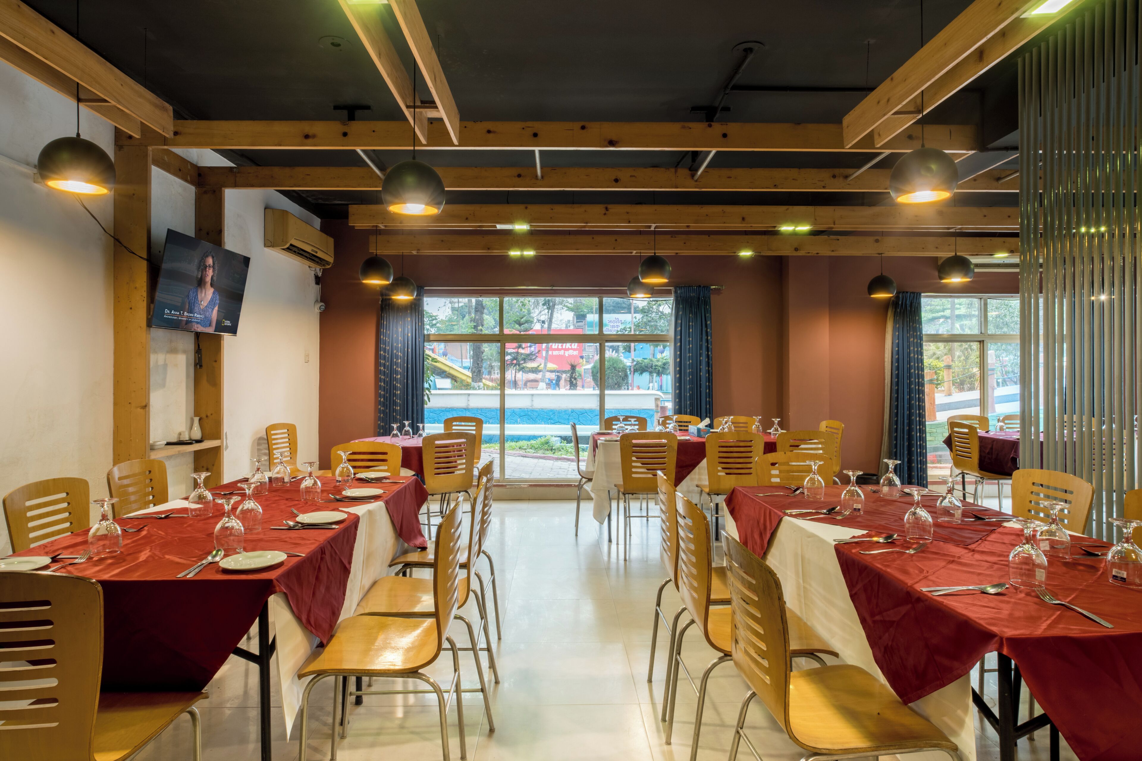Un restaurant amb diverses taules preparades per dinar, un televisor a la paret i grans finestres amb cortines.