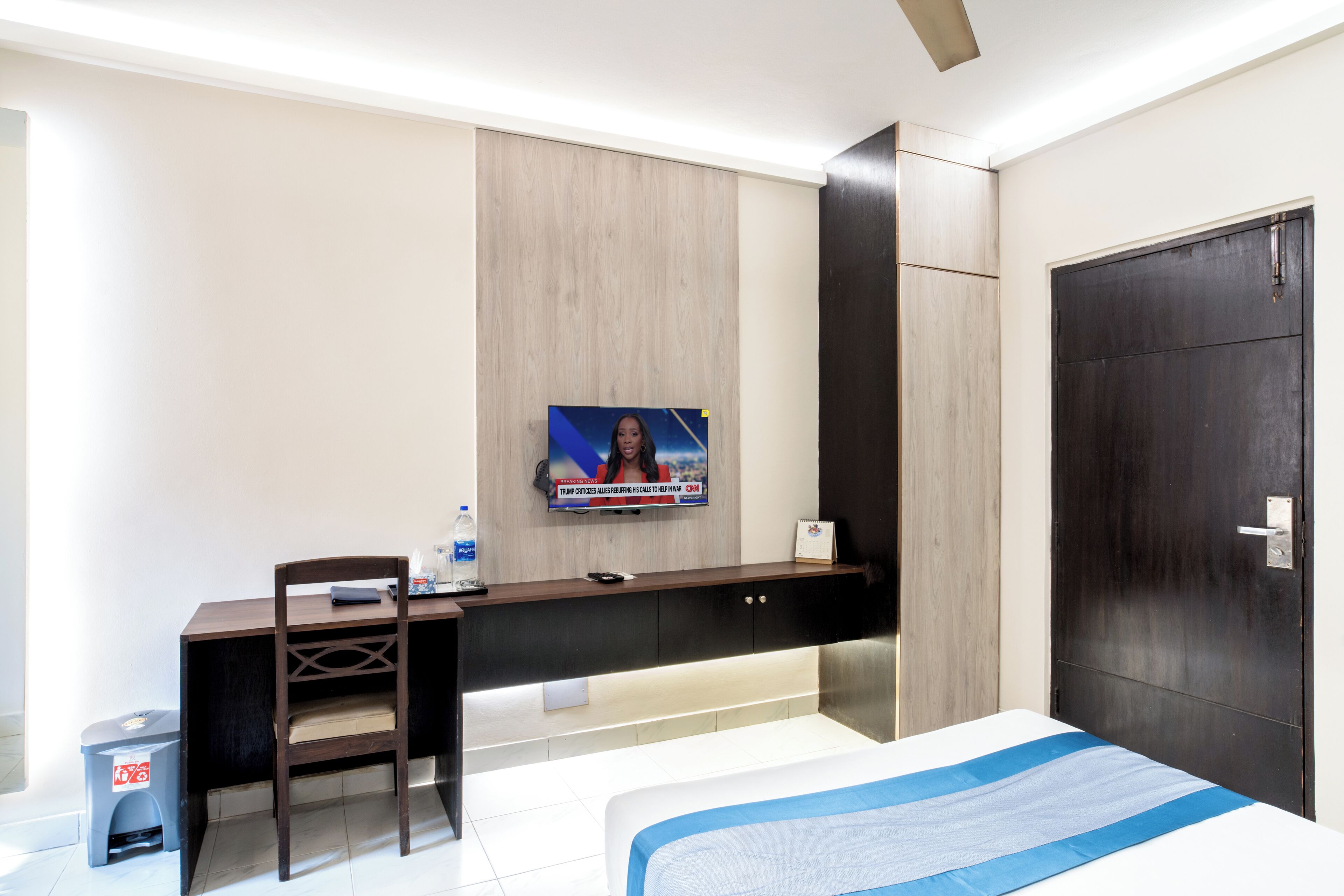 Habitación de hotel con cama, escritorio, silla, televisor montado en la pared y una puerta.