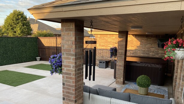 A covered outdoor patio with a brick pillar, a seating area with cushions, a small table, and hanging flower baskets. - Spark Getaways: The Golden Lotus Spa, Sauna, EV Charging (Lubbock)