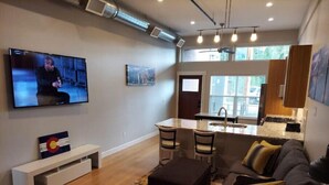 A modern living room with a TV, seating area, and a kitchen in the background.