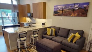 A modern kitchen with a central island, bar stools, a grey sofa with yellow cushions, and a patterned rug.