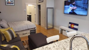 A modern hotel room with a bed, a seating area with a table and chairs, a TV mounted on the wall, and a bathroom visible through an open door.
