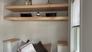 Quarto com cama, duas mesas de cabeceira, janela com persianas e prateleira sobre a cama.