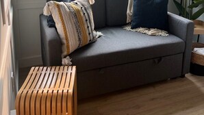Uma sala de estar moderna com um sofá cinzento, um banco de madeira e uma grande janela com vista para árvores e uma estrada.