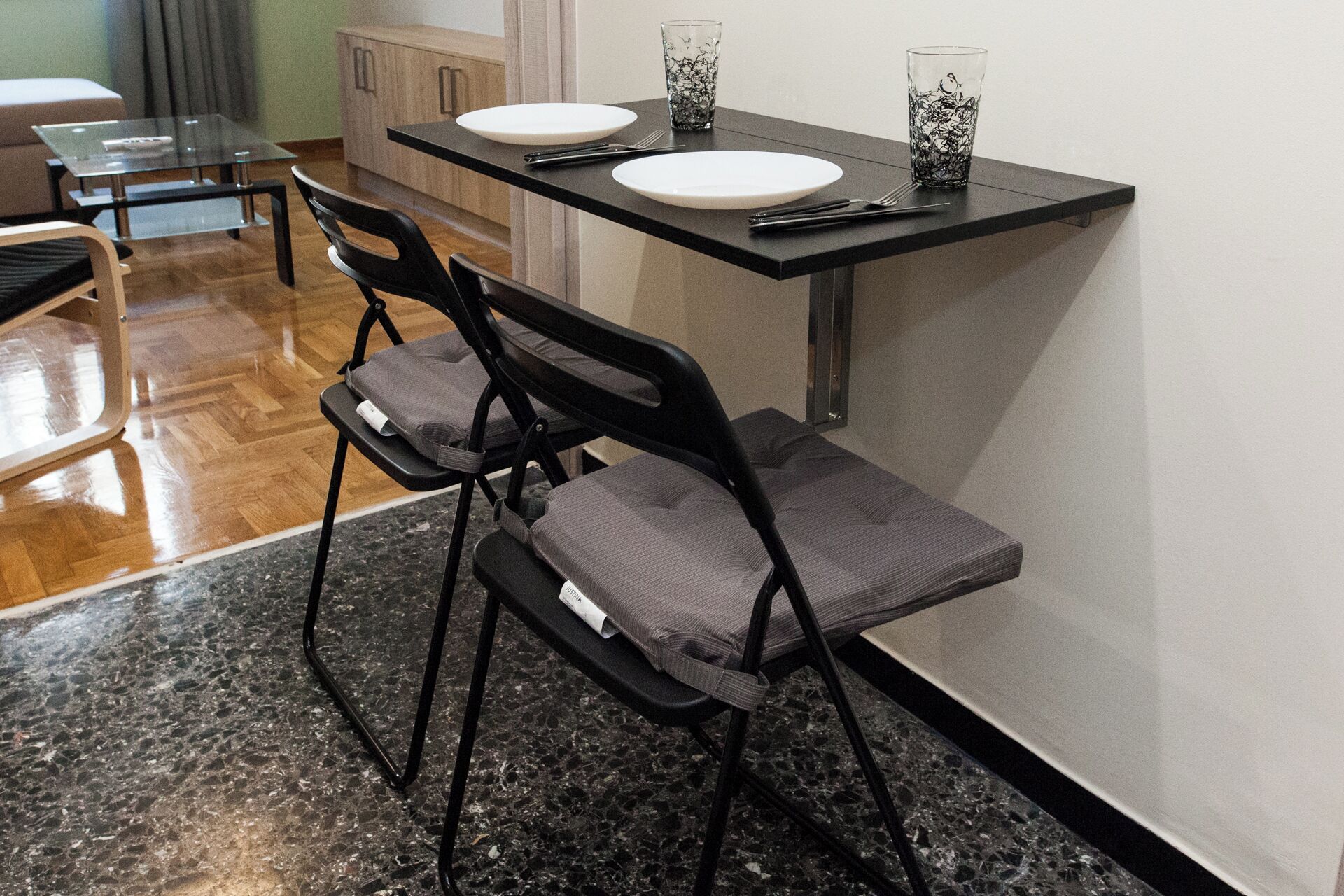 A dining area with two black chairs, a glass table, and a black countertop with a white plate and glass.