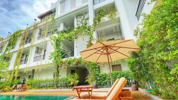Zona junto a la piscina con sillones de madera y una sombrilla, rodeada de exuberante vegetación y un edificio blanco con balcones.