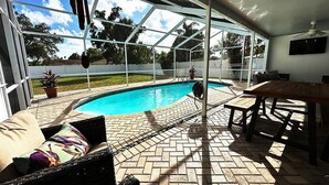 A covered outdoor area with a swimming pool, a wooden table, and a cushioned seating area.