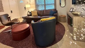 A modern living room with a brown leather sofa, a blue armchair, and a red ottoman. There is a small side table with a lamp, a window with blinds, and a mirror on the wall.