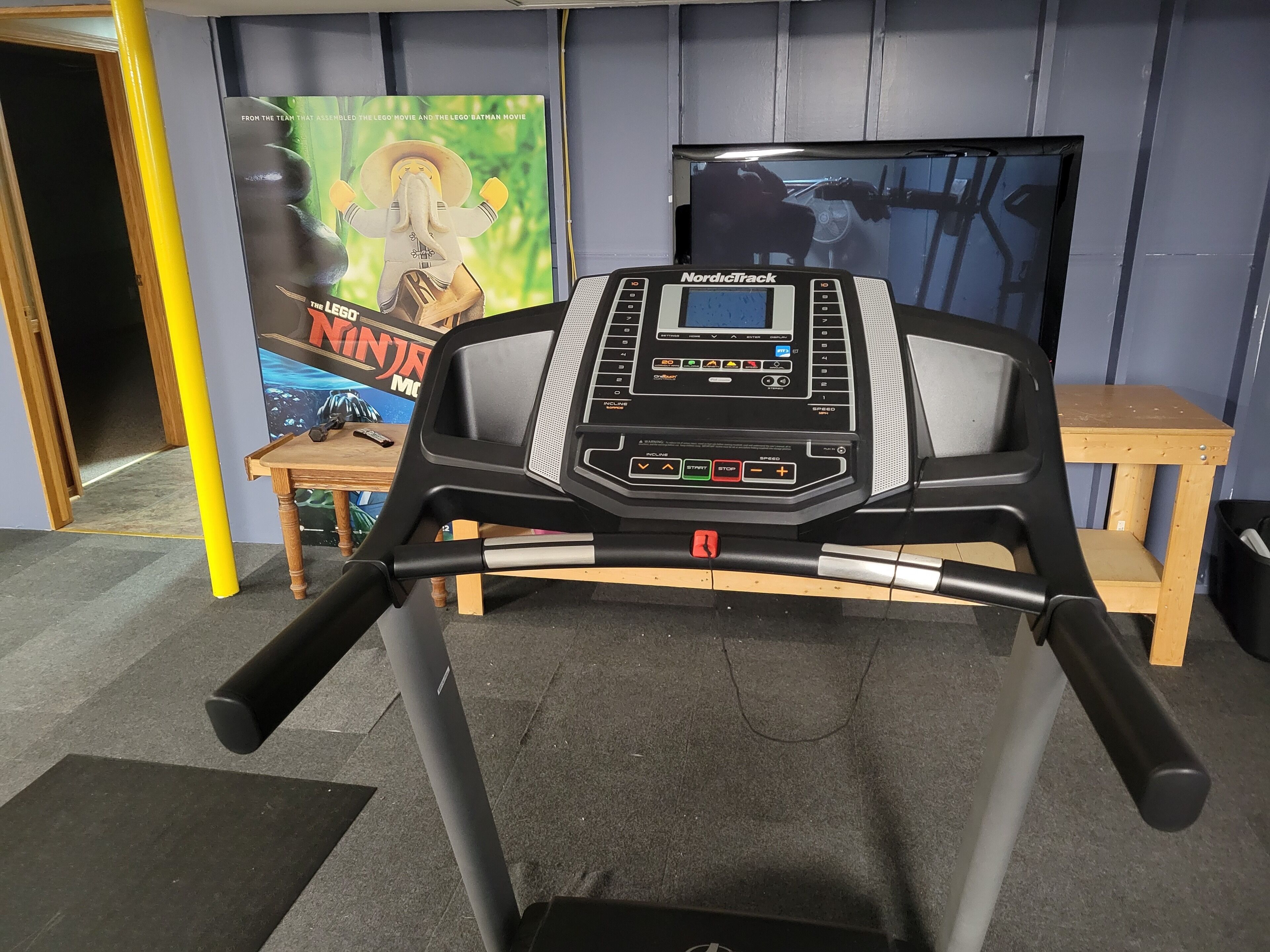 Un tapis de course NordicTrack doté d’un écran numérique, placé dans une pièce où l’on peut voir une affiche d’un film LEGO et une table en bois.