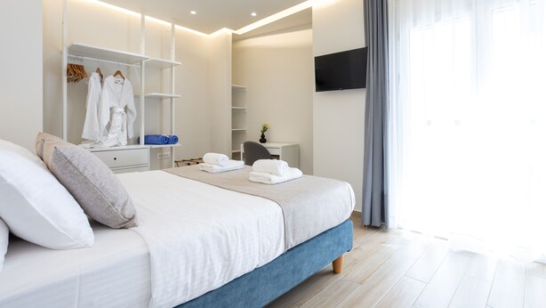 Una camera d'albergo moderna con un letto rifatto, un armadio con accappatoi, una scrivania con una sedia e una TV montata a parete.